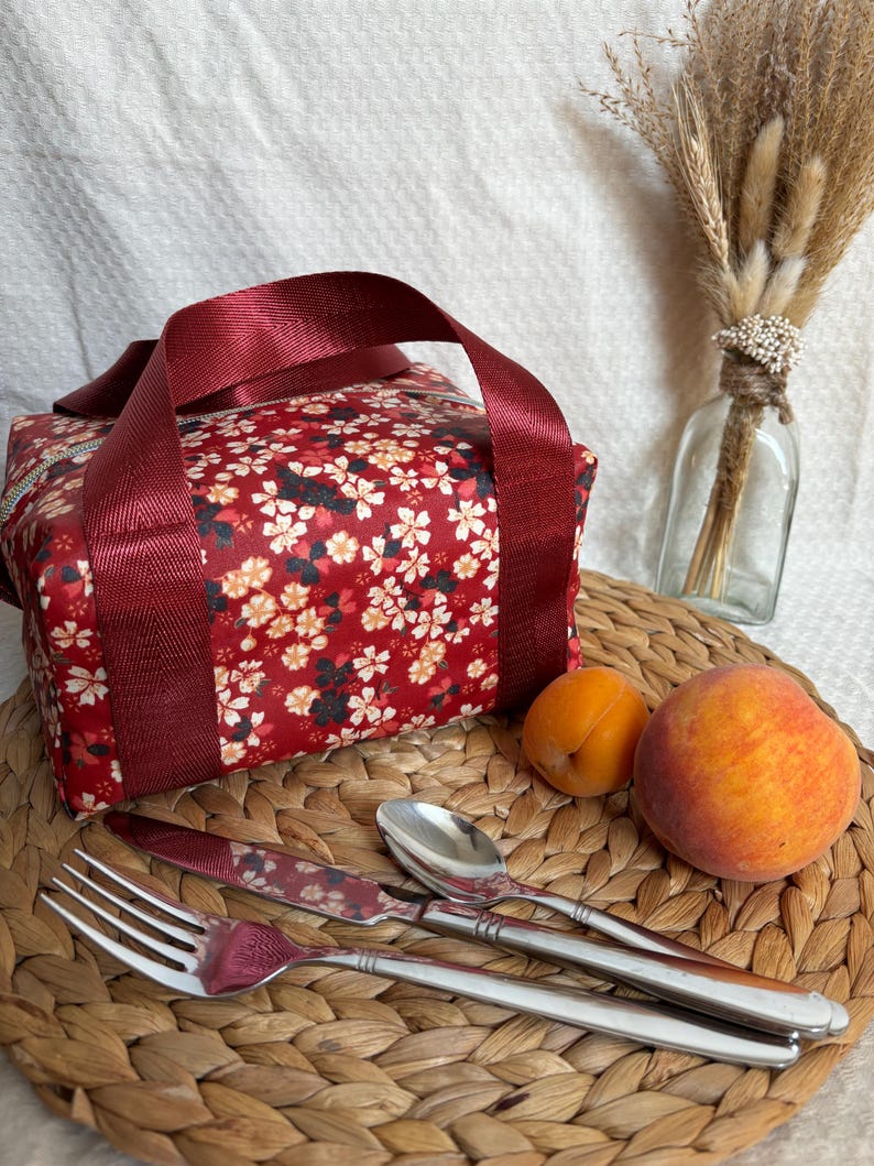 May include: A red lunch bag with a floral print and burgundy straps. The bag is sitting on a woven placemat with a fork, knife, spoon, and two peaches.