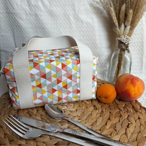 May include: A white lunch bag with a geometric pattern of pink, yellow, blue, and orange triangles. The bag has two white straps and is sitting on a woven wicker surface with a fork, knife, and spoon.