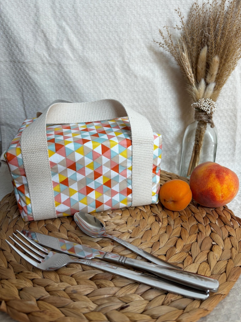 May include: A white lunch bag with a geometric pattern of pink, yellow, blue, and orange triangles. The bag has two white straps and is sitting on a woven wicker surface with a fork, knife, and spoon.