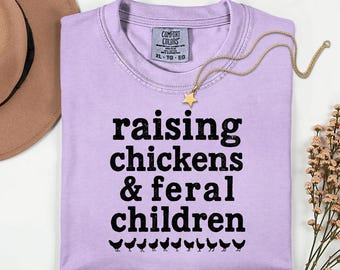 May include: A light purple t-shirt with the text "raising chickens & feral children" in black. A gold necklace with a star pendant rests on the shirt. A brown hat and flowers are in the background.