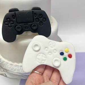 May include: Two fondant video game controller decorations, one black and one white, sit atop a white cake. The white controller has colourful buttons. The black controller is detailed with buttons and directional pads.