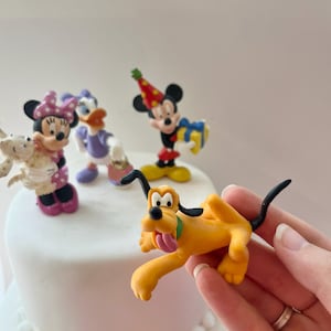May include: A selection of Disney character figurines, including Mickey Mouse, Minnie Mouse, Daisy Duck, and Pluto. Mickey wears a party hat and holds a gift. Pluto is yellow with a green collar and pink tongue.