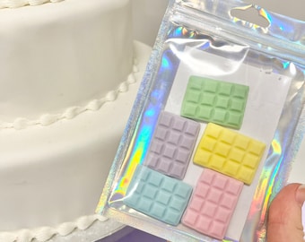 May include: A clear, iridescent resealable pouch containing five small, rectangular, chocolate-bar-shaped items in pastel green, lavender, yellow, light blue, and pink. The items feature a grid pattern on their surface, resembling segmented chocolate bars.
