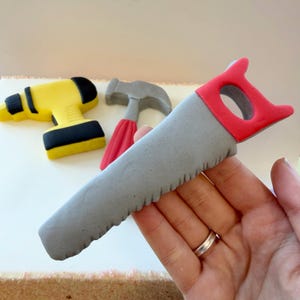 May include: A selection of toy tools, including a grey saw with a red handle, a yellow and black drill, and a grey hammer with a red handle. The tools are designed to look like miniature versions of real tools.