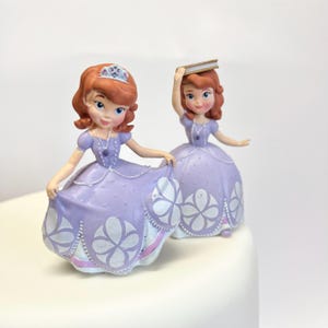 May include: Two small figurines of a character with auburn hair wearing a lavender dress with white floral patterns. One figurine wears a silver tiara and holds the skirt, while the other figurine holds a small rectangular book above its head.