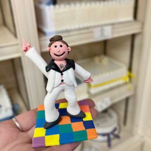 May include: A white fondant figure of a person wearing a white suit with a black shirt and black shoes, dancing on a colourful square dance floor.