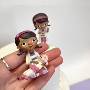 May include: Two small plastic figurines of a cartoon doctor, one holding a small white animal. The doctor figurines are wearing white coats with purple and blue stripes, pink trousers, and pink headbands.