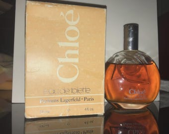 Könnte beinhalten: Eine Vintage-Flasche Chloé Eau de Toilette Parfüm mit einer kugelförmigen Glasflasche und einem silbernen Verschluss. Die Parfümflüssigkeit ist bernsteinfarben. Die Box ist beige mit dem Chloé-Logo und Text.