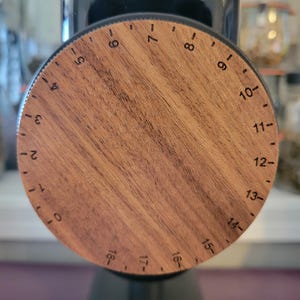 May include: A close-up of a coffee grinder with a wooden dial. The dial is circular with numbers from 0 to 18, and is made of a light brown wood. The grinder is black.