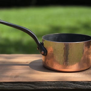 May include: A round copper saucepan with a hammered exterior finish and a long, dark, curved metal handle attached with two rivets. The interior of the pan is dark, and the exterior surface reflects light across its textured, metallic surface.