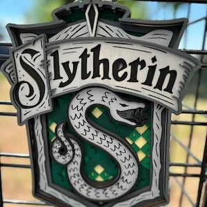 May include: A layered wooden Slytherin house crest, featuring a green and silver serpent on a green and yellow background. The word "Slytherin" is displayed in black lettering on a banner. The crest is framed in gray and black.