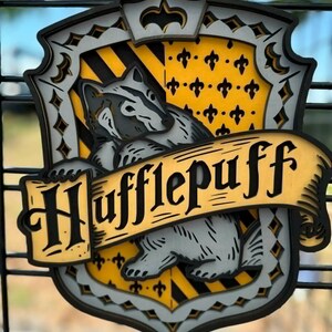 May include: A layered wooden wall plaque featuring the Hufflepuff crest. The design includes a central badger illustration, a yellow and black striped background with fleur-de-lis patterns, and a banner across the front with the text Hufflepuff.