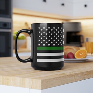 Puede incluir: Taza de café negra con un diseño de bandera estadounidense blanca y una fina franja verde en el medio. La taza está sobre una superficie de madera.