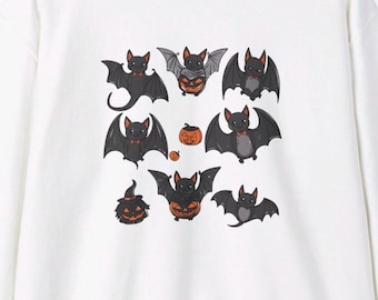 Könnte beinhalten: Weißes Sweatshirt mit einem Halloween-Motiv. Das Design zeigt Cartoon-Fledermäuse mit schwarzen Flügeln und orangefarbenen Fliegen, sowie Kürbislaternen. Die Fledermäuse sind in verschiedenen Posen, was einen verspielten und festlichen Look ergibt.