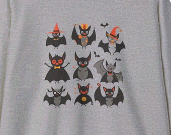 Könnte beinhalten: Ein graues Sweatshirt mit einem Raster von neun Cartoon-Fledermäusen in verschiedenen Posen und Kostümen. Die Fledermäuse sind hauptsächlich schwarz und grau, mit orangefarbenen und gelben Akzenten. Halloween-Thema.