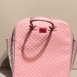 Könnte beinhalten: Rosa gesteppter Rucksack mit oberem Griff und Seitentaschen. Die Tasche hat ein Rautenmuster und einen rechteckigen Aufnäher mit den Buchstaben "HS". Der Rucksack hat einen Reißverschluss.