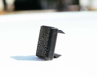 Peut inclure: Un petit objet rectangulaire noir avec une surface texturée présentant un motif topographique en relief composé de lignes concentriques. L'article possède une face avant plate et deux ergots triangulaires saillants à l'arrière.
