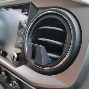 Peut inclure: Un support de t&eacute;l&eacute;phone noir imprim&eacute; en 3D pour la ventilation d'une voiture, install&eacute; sur le tableau de bord d'un v&eacute;hicule. Le support est con&ccedil;u pour s'ins&eacute;rer dans la grille d'a&eacute;ration et maintenir solidement un smartphone. Le tableau de bord est gris avec une garniture argent&eacute;e autour de la ventilation.
