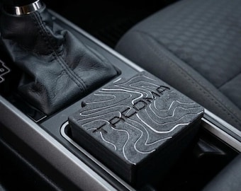 Peut inclure: Un insert de porte-gobelet de voiture carré noir avec un motif de carte topographique et le mot TACOMA en relief sur la surface. L'insert est placé dans la console centrale à côté d'un soufflet de levier de vitesse en cuir noir.