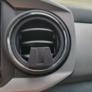 Peut inclure: Un support de t&eacute;l&eacute;phone noir install&eacute; dans une bouche d'a&eacute;ration de voiture. Le support est con&ccedil;u pour maintenir un t&eacute;l&eacute;phone en place. Le tableau de bord environnant est gris et noir, avec une bouche d'a&eacute;ration circulaire et une garniture argent&eacute;e.