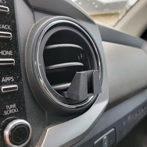 Peut inclure: Un support de t&eacute;l&eacute;phone noir imprim&eacute; en 3D pour la ventilation d'une voiture, install&eacute; dans une bouche d'a&eacute;ration du tableau de bord du v&eacute;hicule. Le support est con&ccedil;u pour maintenir solidement un smartphone. Le tableau de bord est gris et noir.