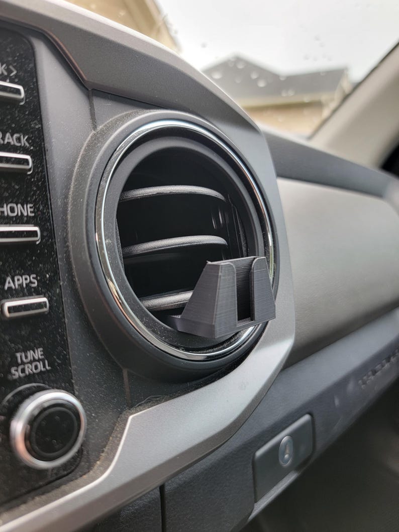 Peut inclure: Un support de t&eacute;l&eacute;phone noir imprim&eacute; en 3D pour la ventilation d'une voiture, install&eacute; dans une bouche d'a&eacute;ration du tableau de bord du v&eacute;hicule. Le support est con&ccedil;u pour maintenir solidement un smartphone. Le tableau de bord est gris et noir.