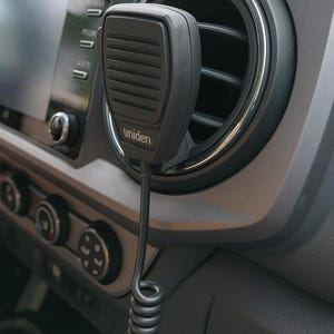 May include: A black Uniden microphone with a coiled cord is mounted on a vehicle's dashboard. The microphone is positioned near an air vent and the vehicle's infotainment screen. The dashboard is gray.