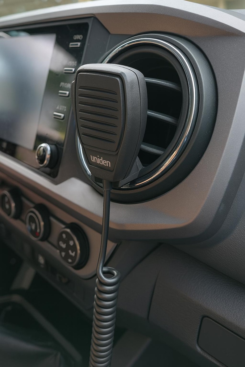 Peut inclure: Un microphone Uniden noir avec un cordon enroul&eacute; est mont&eacute; sur le tableau de bord d'un v&eacute;hicule. Le microphone est positionn&eacute; pr&egrave;s d'une bouche d'a&eacute;ration et de l'&eacute;cran d'infodivertissement du v&eacute;hicule. Le tableau de bord est gris.
