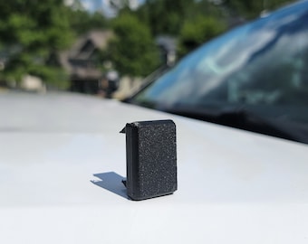 Peut inclure: Une pièce en plastique noire et rectangulaire avec une surface texturée. La pièce a une petite extension angulaire sur un côté et repose sur une surface blanche. L'arrière-plan est flou, montrant des arbres et un véhicule.