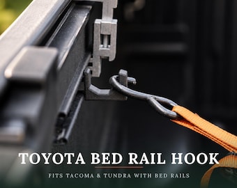 Peut inclure: Un crochet de rail de benne en métal noir fixé sur un système de rail de camion, doté d'une forme incurvée retenant une sangle en nylon orange vif. Le texte TOYOTA BED RAIL HOOK FITS TACOMA & TUNDRA WITH BED RAILS est inscrit en bas.