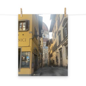 Puede incluir: Fotografía de una calle estrecha en Florencia, Italia, con edificios en tonos amarillos y beige. La cúpula del Duomo es visible a lo lejos. La imagen está impresa en papel y sujeta con pinzas de madera.