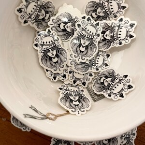 Könnte beinhalten: Eine Schüssel gefüllt mit schwarzen und weißen Aufklebern, die eine Frau mit langen Haaren zeigen, die eine Krone aus Pilzen trägt. Die Aufkleber befinden sich auf einem weißen Hintergrund.