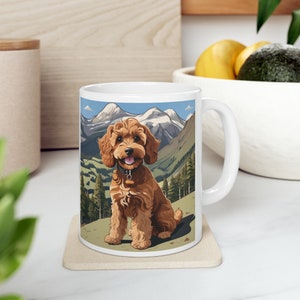 Könnte beinhalten: Weiße Keramiktasse mit einer braunen und weißen Illustration eines Hundes, der in einer Berglandschaft sitzt. Der Hund ist ein brauner Pudel mit einem schwarzen Halsband und einem braunen Anhänger. Die Berge sind im Hintergrund und mit Schnee bedeckt. Die Tasse hat einen weißen Henkel.