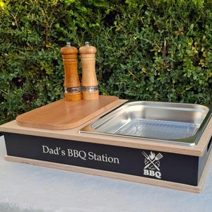 Könnte beinhalten: Eine hölzerne BBQ-Station mit einem Edelstahlbehälter und einem Holzschneidebrett. Auf der schwarzen Frontplatte steht "Dad's BBQ Station" mit einem BBQ-Logo. Zwei hölzerne Salz- und Pfeffermühlen stehen auf dem Schneidebrett.