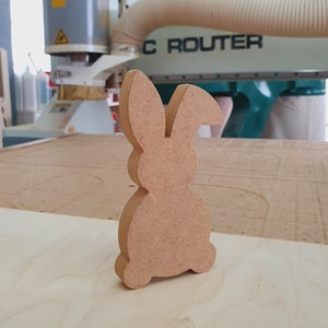 May include: A wooden rabbit cutout, unpainted, on a light brown wood surface. The rabbit is in the foreground, with a CNC router in the background.