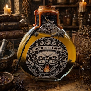 Op de afbeelding: Een ronde glazen fles gevuld met een gele vloeistof, met een kurk afgesloten met een donkere substantie en een mot-hanger. Het etiket luidt "Moth to a Flame" met een mot-illustratie en een vlam. De fles wordt omringd door boeken en kaarsen.