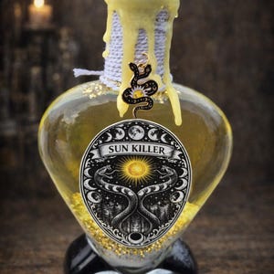 May include: A heart-shaped glass bottle filled with yellow liquid, featuring a cork stopper and a decorative label with the text "SUN KILLER". The bottle is adorned with a snake charm and gold flecks.
