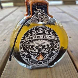 May include: A round glass bottle filled with yellow liquid and black granules. The bottle has a cork stopper sealed with orange wax and a silver moth charm. The label reads "Moth to a Flame" with a moth illustration.