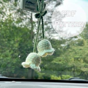 May include: A pair of crocheted, bell-shaped ornaments in cream color, suspended by green yarn. The image includes the text "PDF PATTERN" and "MARIA FLORA" in a white circle, suggesting a craft project. The background is blurred, showing a green, natural environment.
