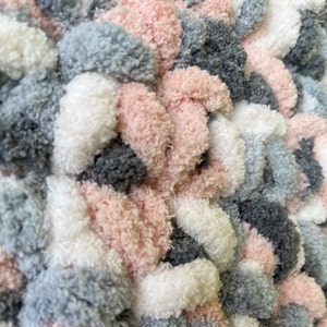 May include: A close-up of a fluffy, textured fabric with a repeating pattern of pink, gray, and white pom-poms. The fabric is soft and plush, and the pom-poms are arranged in a staggered pattern.