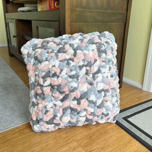 May include: A soft, plush pillow with a textured, bumpy surface. The pillow is made of a mix of pink, white, and gray yarn.