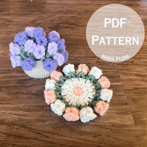 May include: Two crocheted flower patterns, one in purple and one in pink, white, and green. The patterns are on a wooden surface with the text "PDF PATTERN MARIA FLORA" in a white circle.