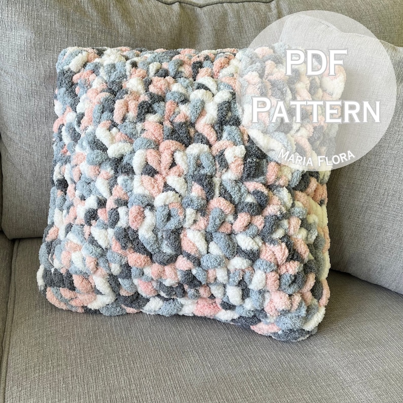 May include: A gray, white, and pink crocheted pillow with a fluffy texture. The pillow is on a gray couch. The text "PDF PATTERN MARIA FLORA" is in a white circle on the right side of the image.