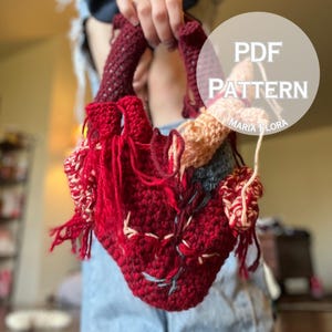 Puede incluir: Un bolso hecho a mano con forma de corazón tejido a ganchillo en tonos rojos, rosas y grises. El bolso presenta costuras intrincadas y detalles de flecos, con el texto "PDF PATTERN MARIA FLORA" en un círculo blanco. El bolso tiene dos asas burdeos.