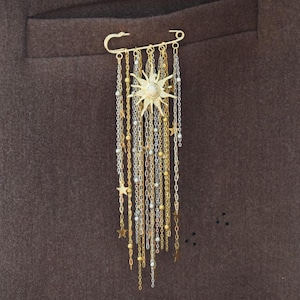 Puede incluir: Broche dorado con diseño de sol, perla central y cadenas colgantes. Las cadenas están adornadas con pequeñas estrellas y cuentas. El broche está sujeto a un imperdible, sobre un fondo de tela marrón.