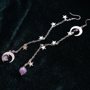 Può includere: Orecchini pendenti color argento con un design celeste. Ogni orecchino ha un ciondolo a forma di luna crescente e stelle, con una catena di stelle. Gli orecchini hanno anche una gemma viola nella parte inferiore e nella luna.