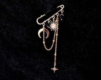 May include: Gold-colored celestial-themed safety pin. The pin features a crescent moon, sun, and star charms, along with dangling chains and beads. The pin is designed with a celestial theme, perfect for adding a touch of cosmic flair to any outfit.