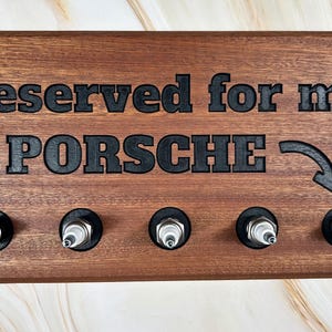 May include: A wooden sign with the text "Reserved for my PORSCHE" and an arrow pointing to the right. The sign has five slots for spark plugs.