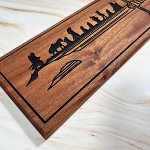 May include: A wooden sign with a black outline and a design of a sword and a line of figures walking towards it. The design is reminiscent of the Lord of the Rings.