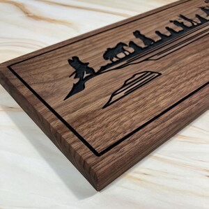 May include: A wooden sign with a black border and a laser-etched design of a group of people walking across a bridge. The design is in a silhouette style.